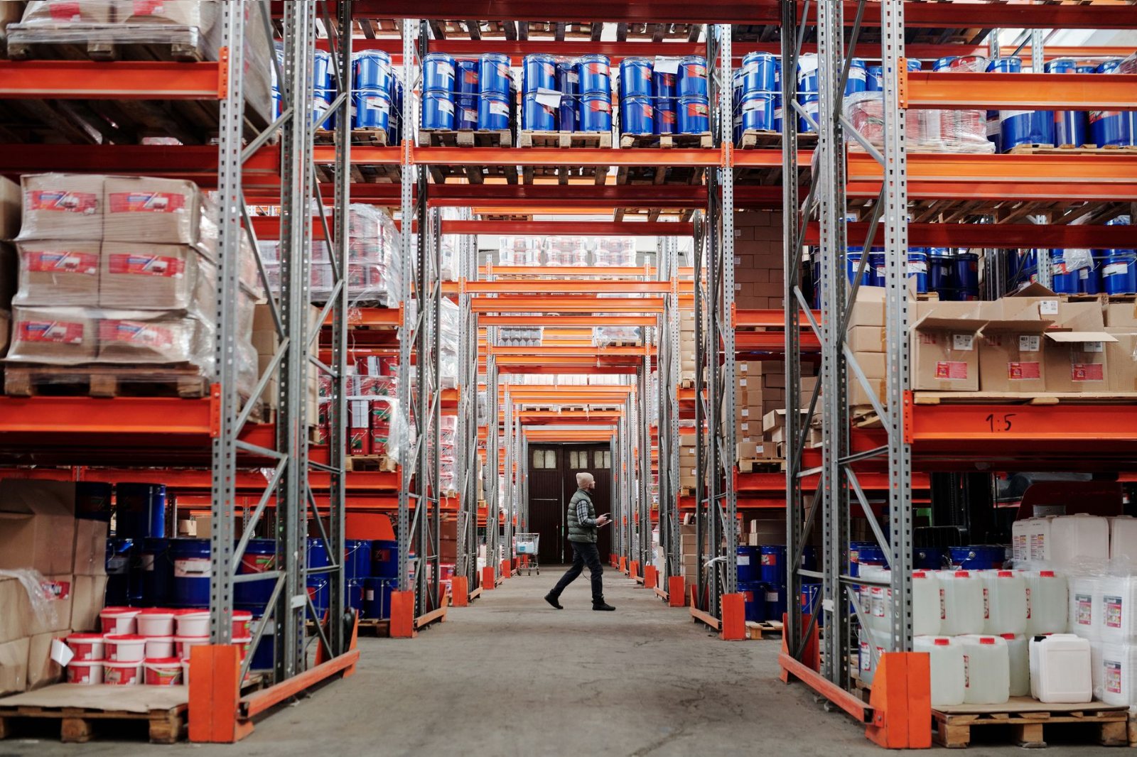 Cartons and shrink-wrapped pallets racked in an orderly warehouse bay, racks filled to the upper level, a pallet jack parked at the end of the marked aisle and yellow floor tape running the lane, a clipboard resting on a stacked pallet