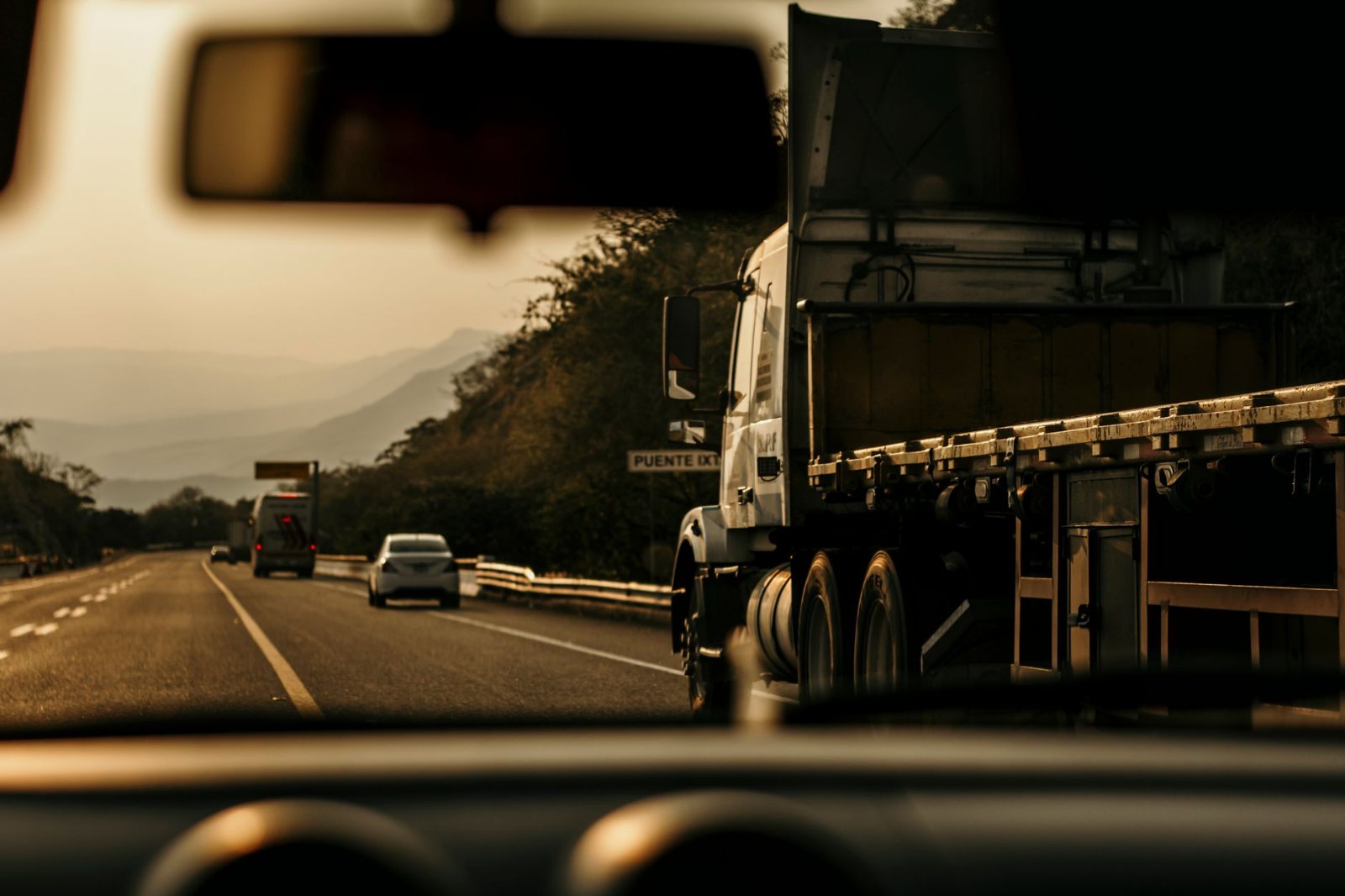 A tractor-trailer at highway speed on an open interstate, framed from behind and slightly alongside in the adjacent lane so no carrier name, DOT number or livery reads; lane lines and low sun ahead, guardrail and open country either side